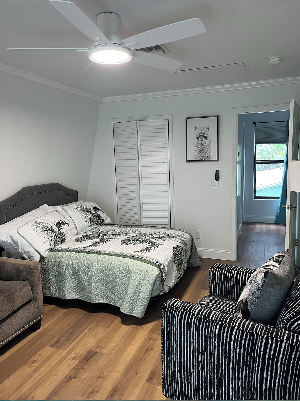 A bedroom with a bed, a chair, a ceiling fan, and a door leading to another room.