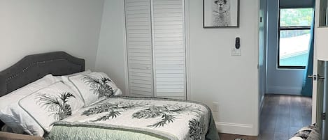 A bedroom with a bed, a chair, a ceiling fan, and a door leading to another room.