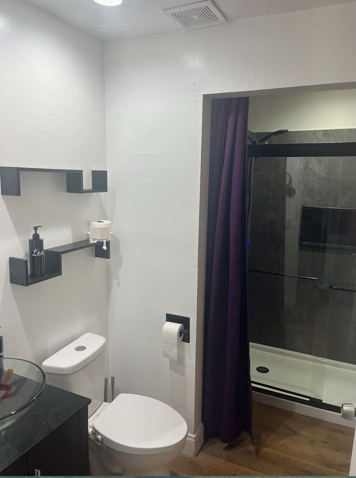 A bathroom with a toilet, a glass shower enclosure, and a shelf with toiletries.