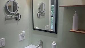 A bathroom with a white sink, a mirror, and a toilet. There are three lights above the mirror and a shelf with a bottle.