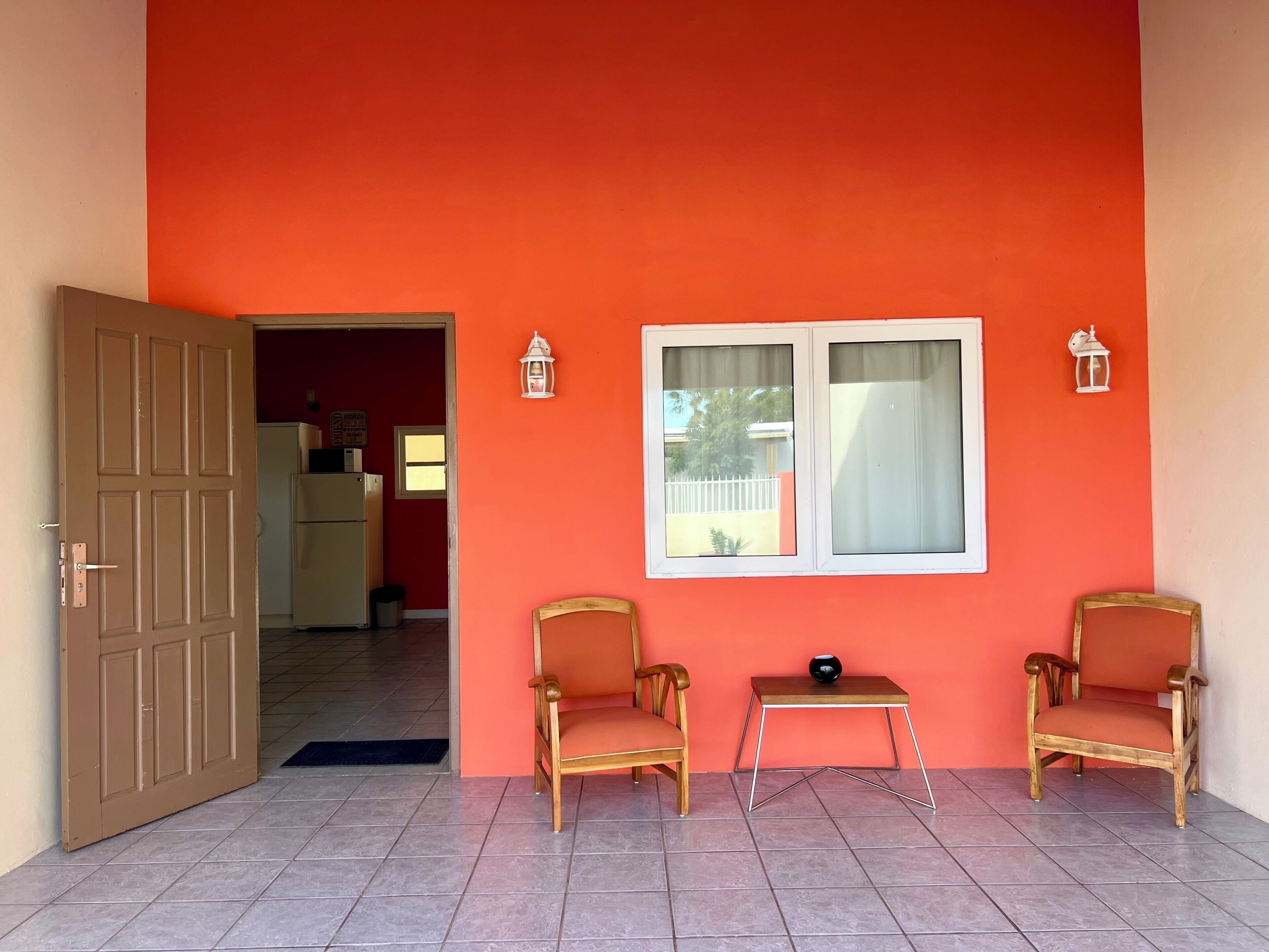 Une pièce avec un mur rouge, deux chaises en bois, une petite table et une porte donnant sur une autre pièce.