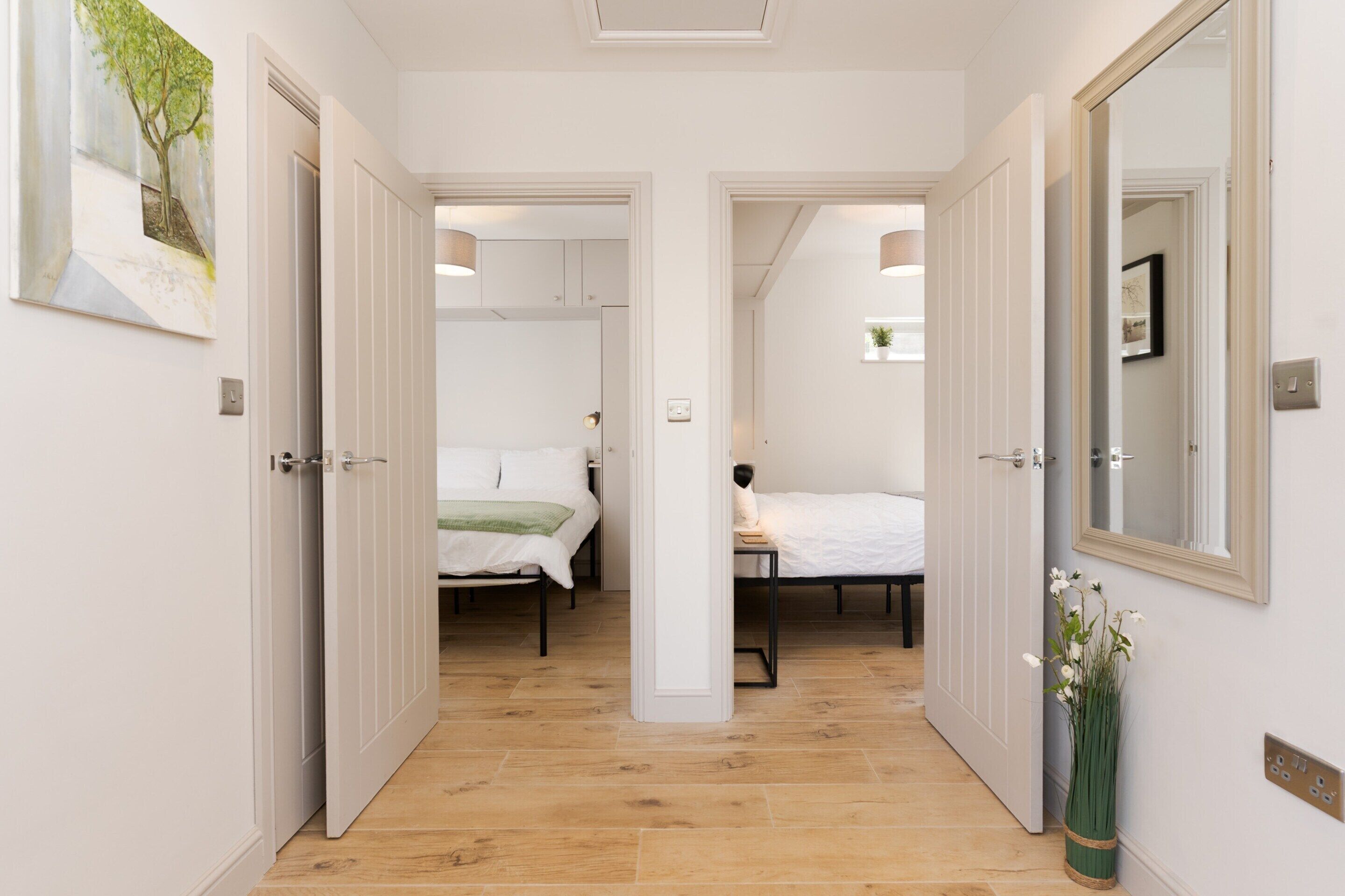 A hallway leading to two bedrooms with wooden flooring, white walls, and a mirror on the right.