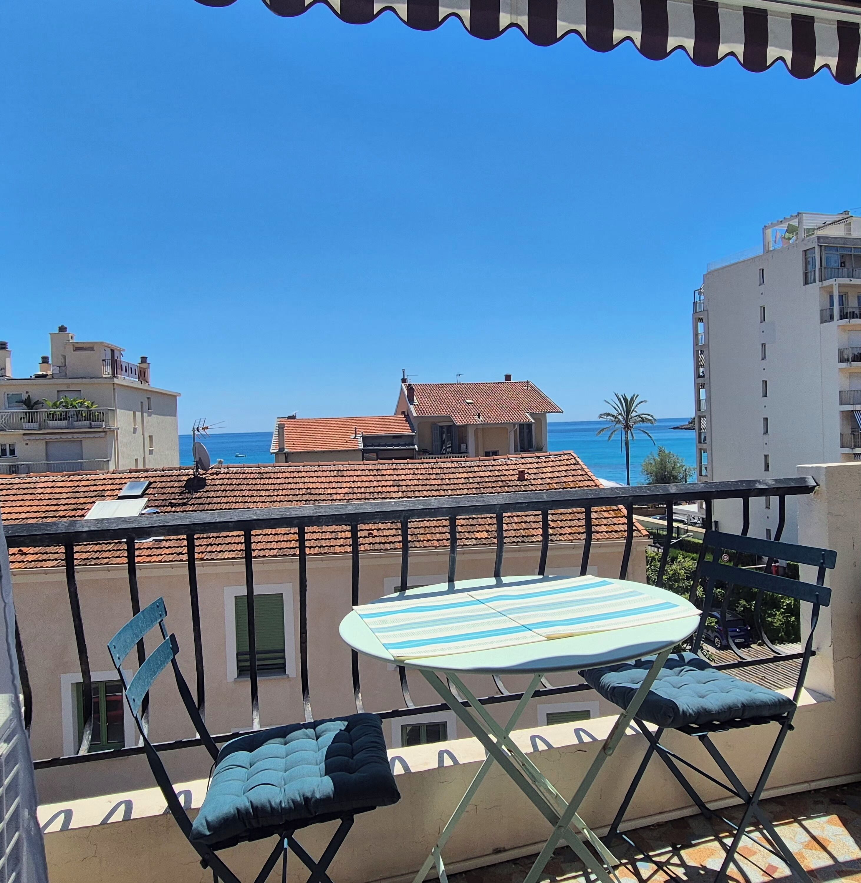 A balcony with a table and two chairs, overlooking a coastal area with buildings and a clear sky.