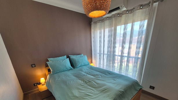 A bedroom with a bed, a window with curtains, a wall-mounted air conditioner, and a hanging light fixture. - New, bright 2-room apartment with ocean views and direct access to the beaches, near Menton and Monaco (Roquebrune-Cap-Martin)