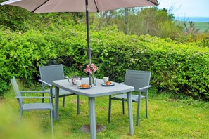 Ein Tisch mit Stühlen im Freien, ein Sonnenschirm und eine gedeckte Tafel mit Teekanne, Tassen und Gebäck.
