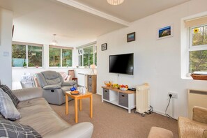 Ein modernes Wohnzimmer mit Sofa, Sessel und Fernseher. Fenster bieten Blick auf Grünflächen.