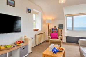 Ein Wohnzimmer mit Flachbildfernseher, Heizkörper, Sessel mit Decke, Tisch mit Teekanne und Croissants sowie Meerblick durch ein Fenster.