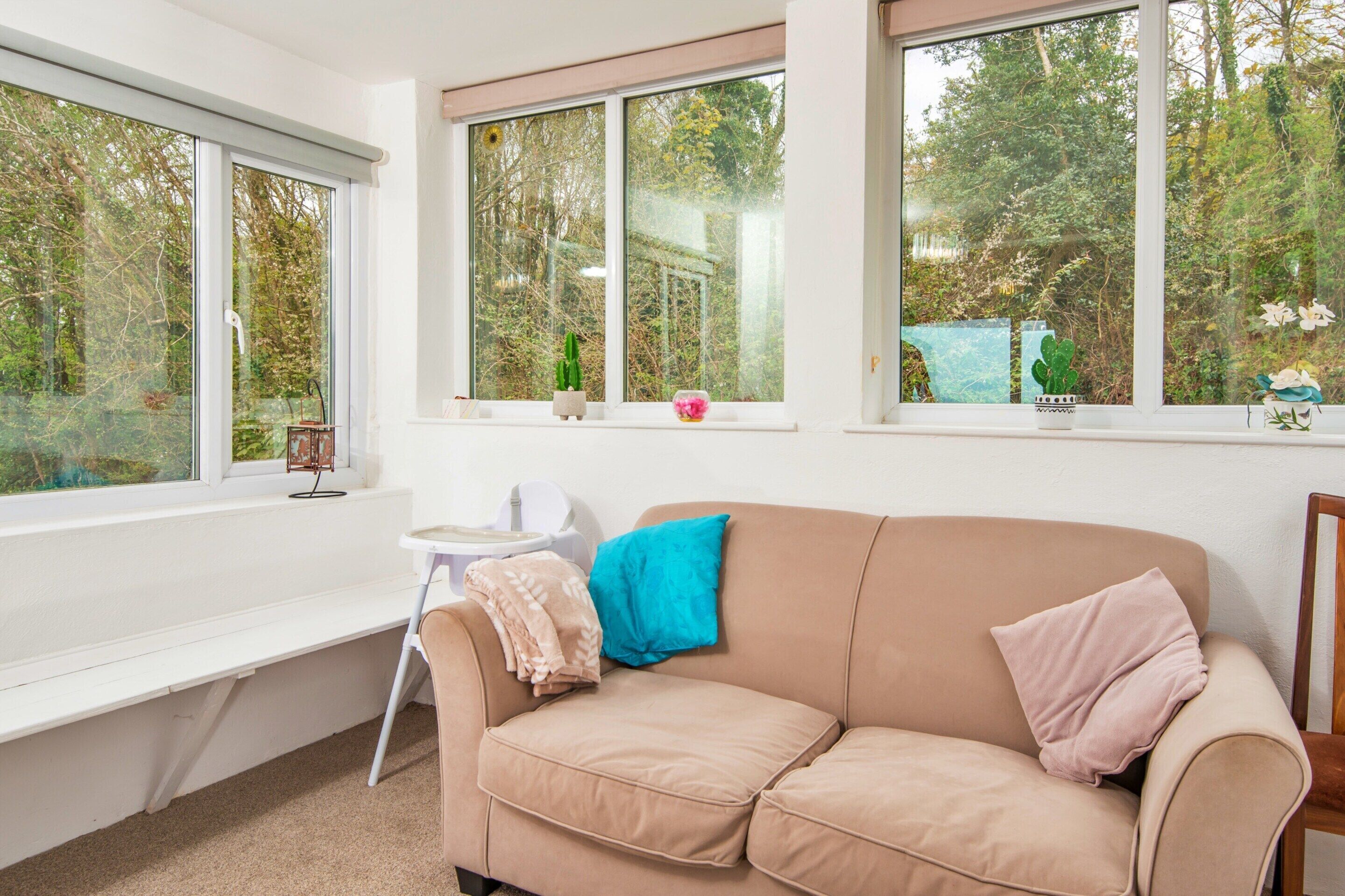 A room with a beige sofa, a small white table, and a bench. There are three windows with views of trees and a garden.