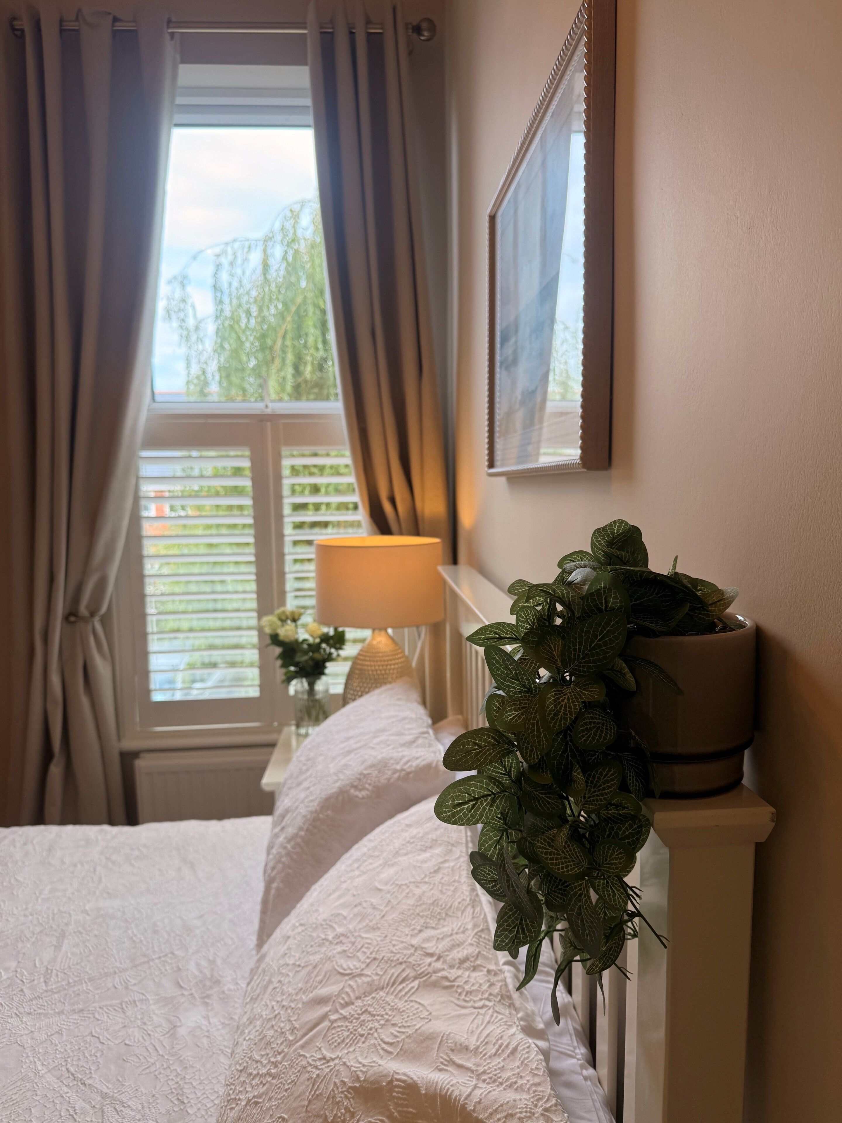 A bedroom with a bed, a bedside lamp, a potted plant, and a window with curtains.