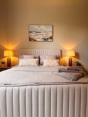A bedroom with a large bed, two bedside lamps, a painting of a beach scene, and a neatly made bed with a folded towel.