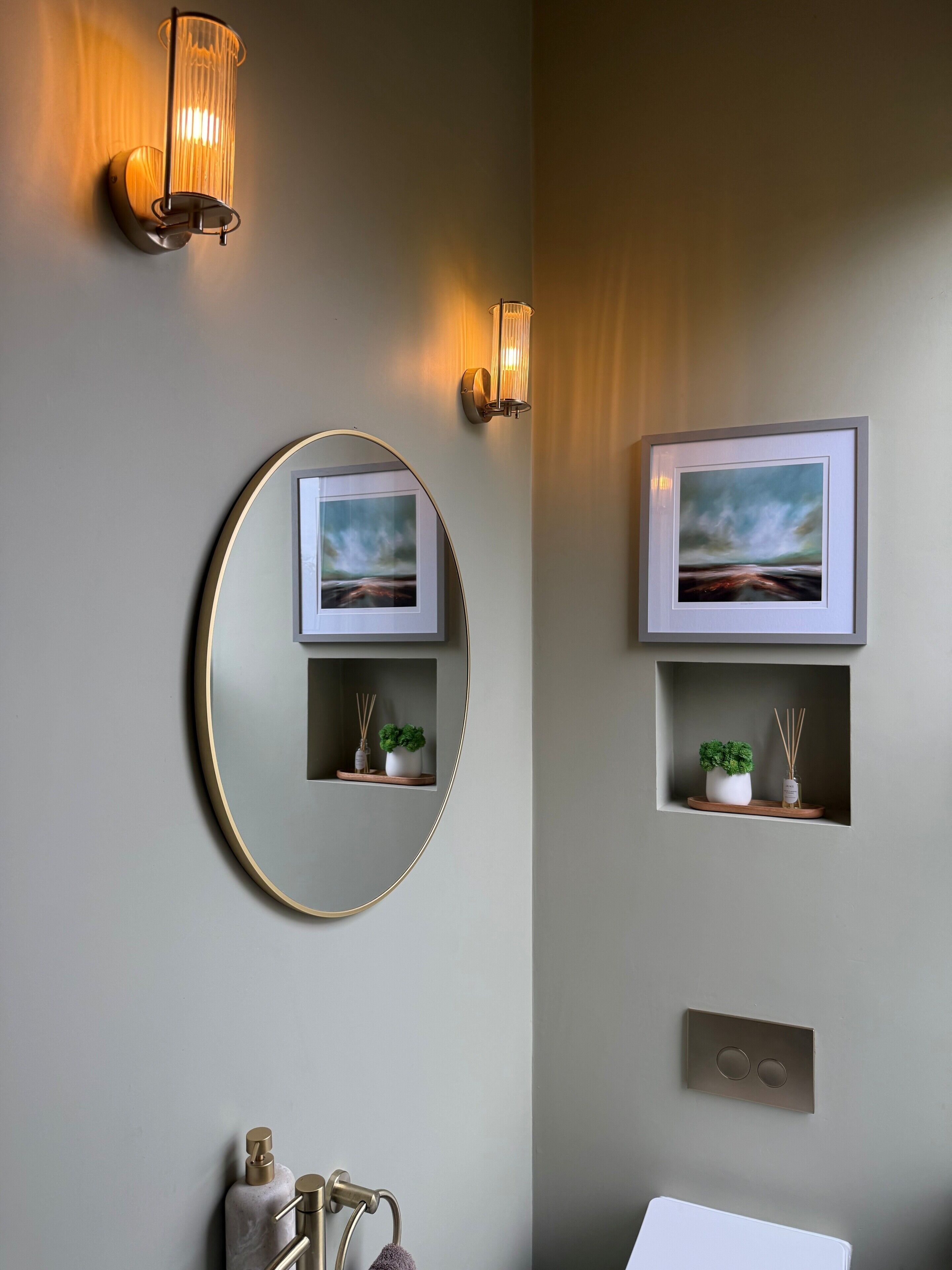 A bathroom with a round mirror, two wall-mounted lights, a framed picture, and a small shelf with a plant and reed diffuser.