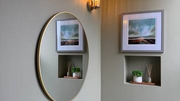 A bathroom with a round mirror, two wall-mounted lights, a framed picture, and a small shelf with a plant and reed diffuser.