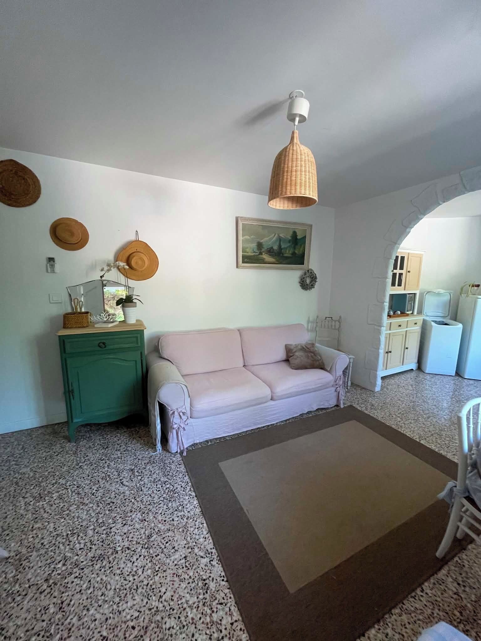 Een woonkamer met een groen kastje, een witte bank, een geweven hanglamp en een lijstwerk schilderij van een landschap aan de muur.