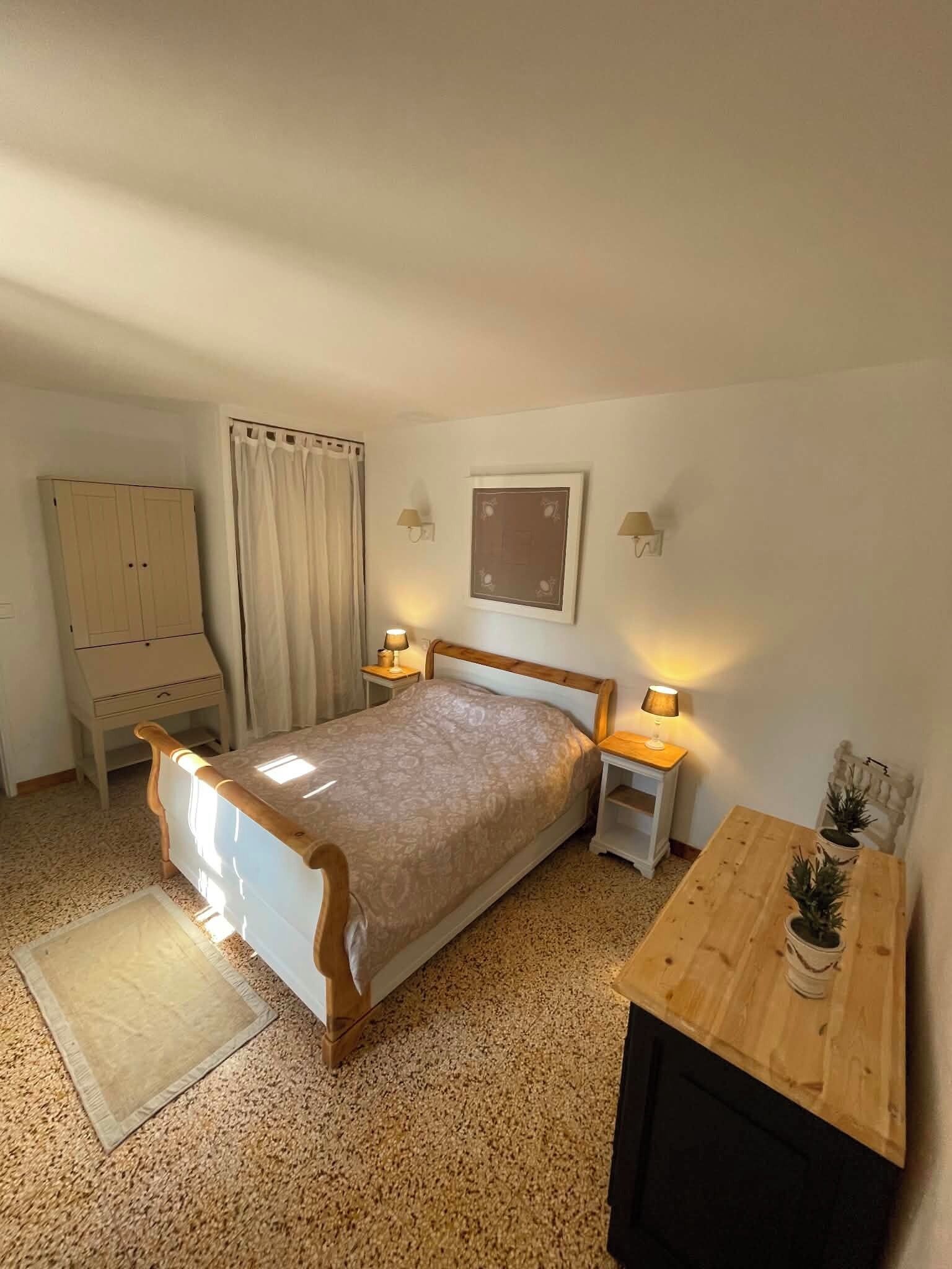 A bedroom with a bed, bedside tables, a wooden nightstand, a desk, a chair, a wardrobe, and a window with curtains.