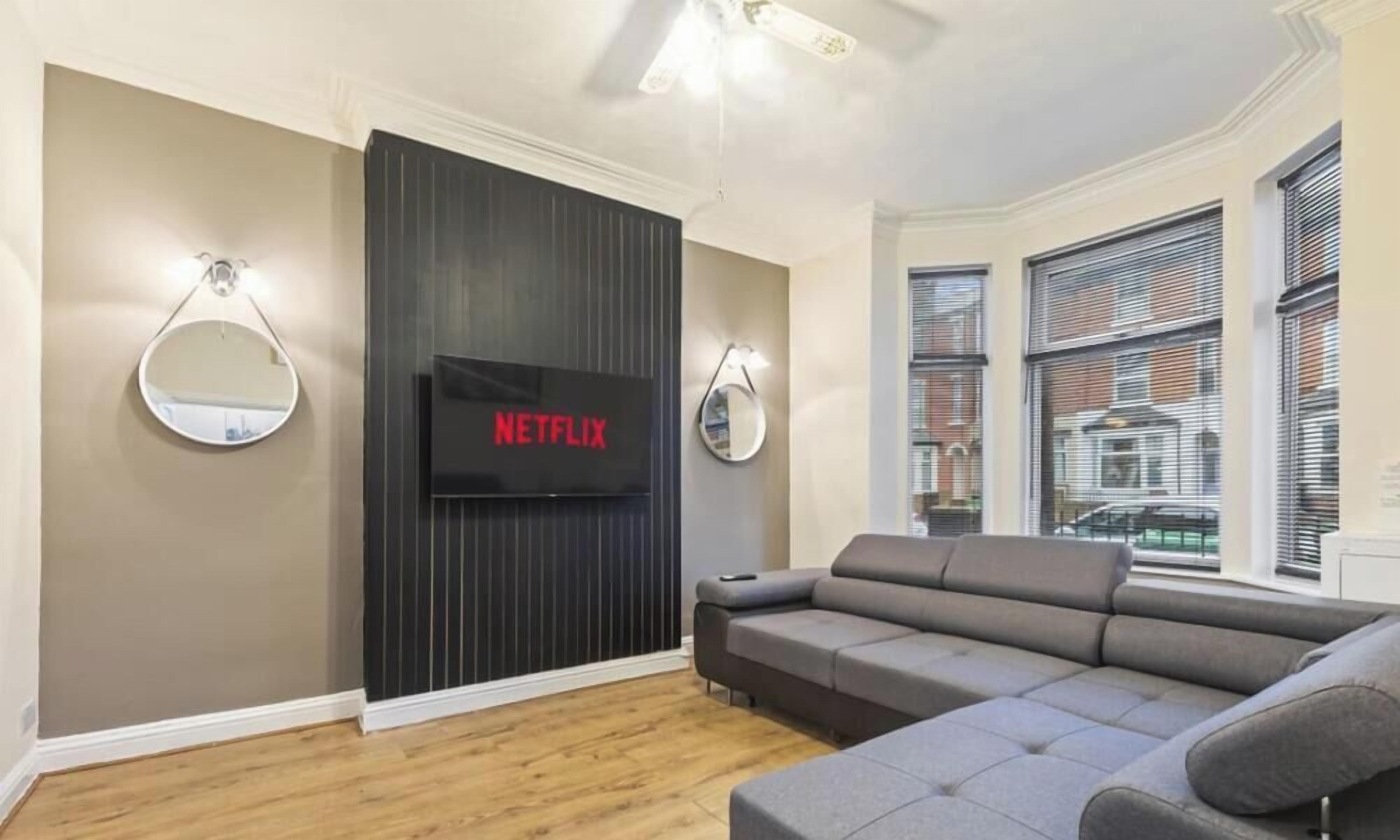 A modern living room with a grey sofa, a flat-screen TV mounted on a dark wall, and two wall-mounted mirrors.