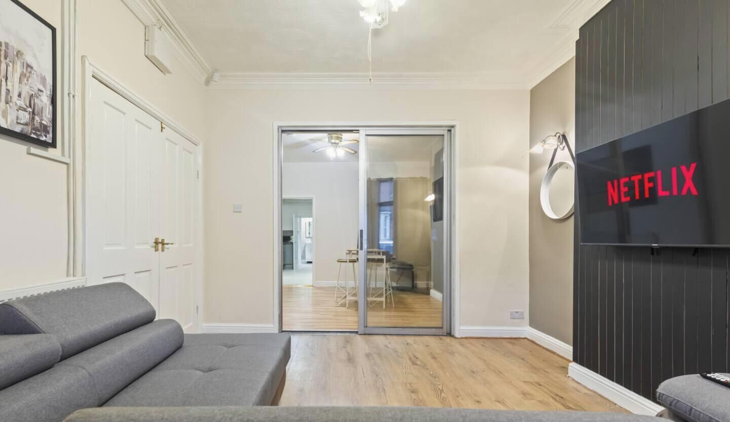 A room with a grey sofa, a sliding glass door leading to a kitchen, and a wall-mounted Netflix sign.