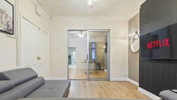 A room with a grey sofa, a sliding glass door leading to a kitchen, and a wall-mounted Netflix sign.