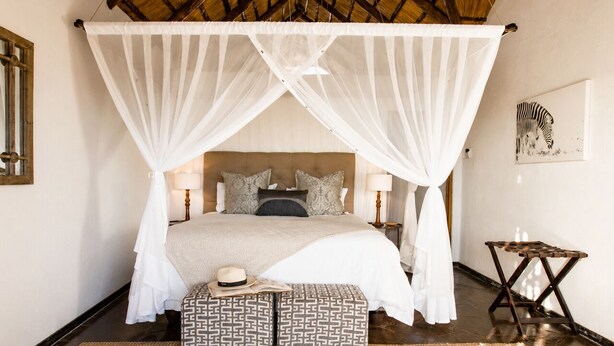 Un dormitorio con cama a dosel, mesitas de noche, un taburete pequeño y una obra de arte con diseño de cebra en la pared.