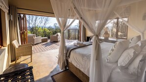 Un dormitorio con cama, cortinas blancas, un balcón con mobiliario de mimbre y vistas a árboles.