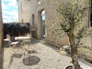 Un bâtiment en pierre doté d’une terrasse en gravier, de sièges extérieurs et d’un arbre en pot.