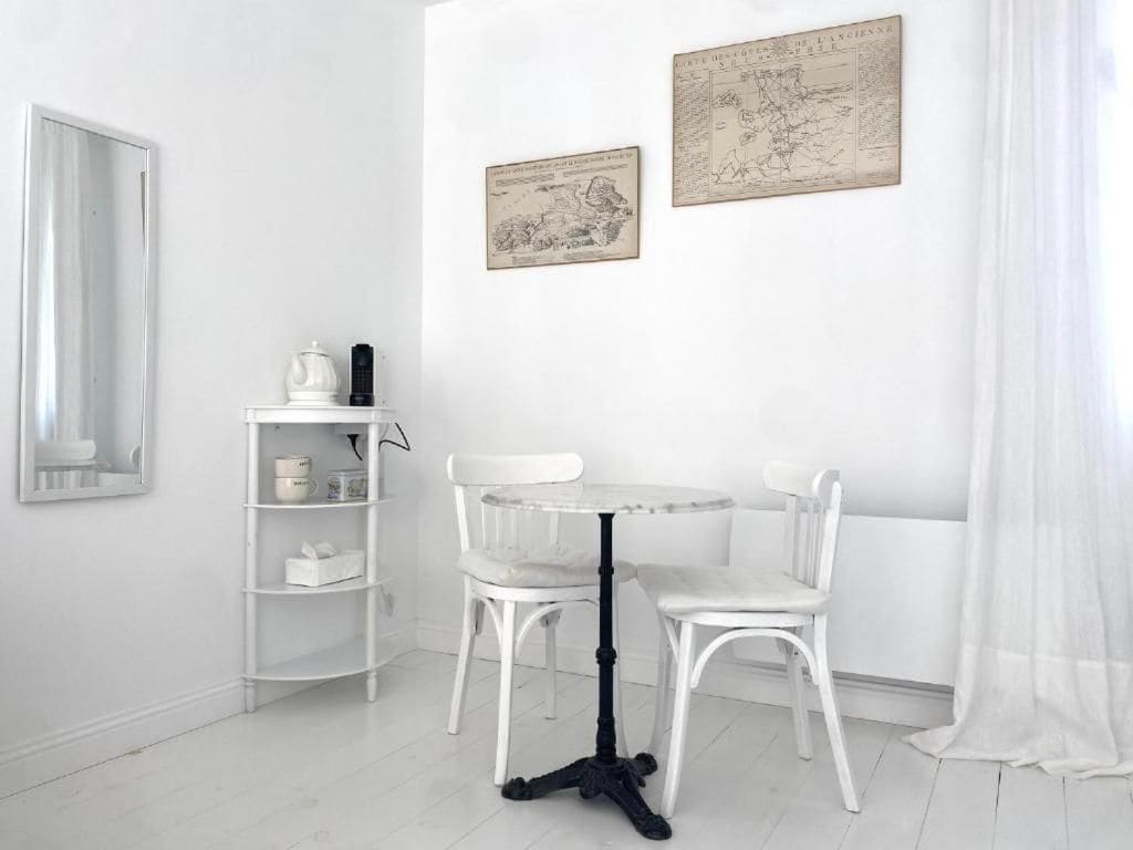 Un coin repas aux murs blancs, avec une table ronde et deux chaises, une étagère blanche où sont posés divers objets, et deux cartes encadrées accrochées au mur.