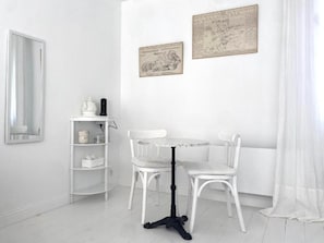 A white dining area with a round table and two chairs, a white shelf with items, and two framed maps on the wall.