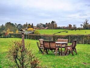 Ein grünes Areal im Freien mit einem Holztisch und Stühlen, umgeben von einem Hecken und einem Baum.