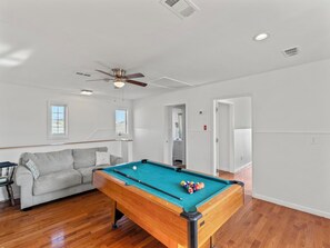 Ein Billardtisch in einem Raum mit Holzböden, einem Deckenventilator und einem grauen Sofa.