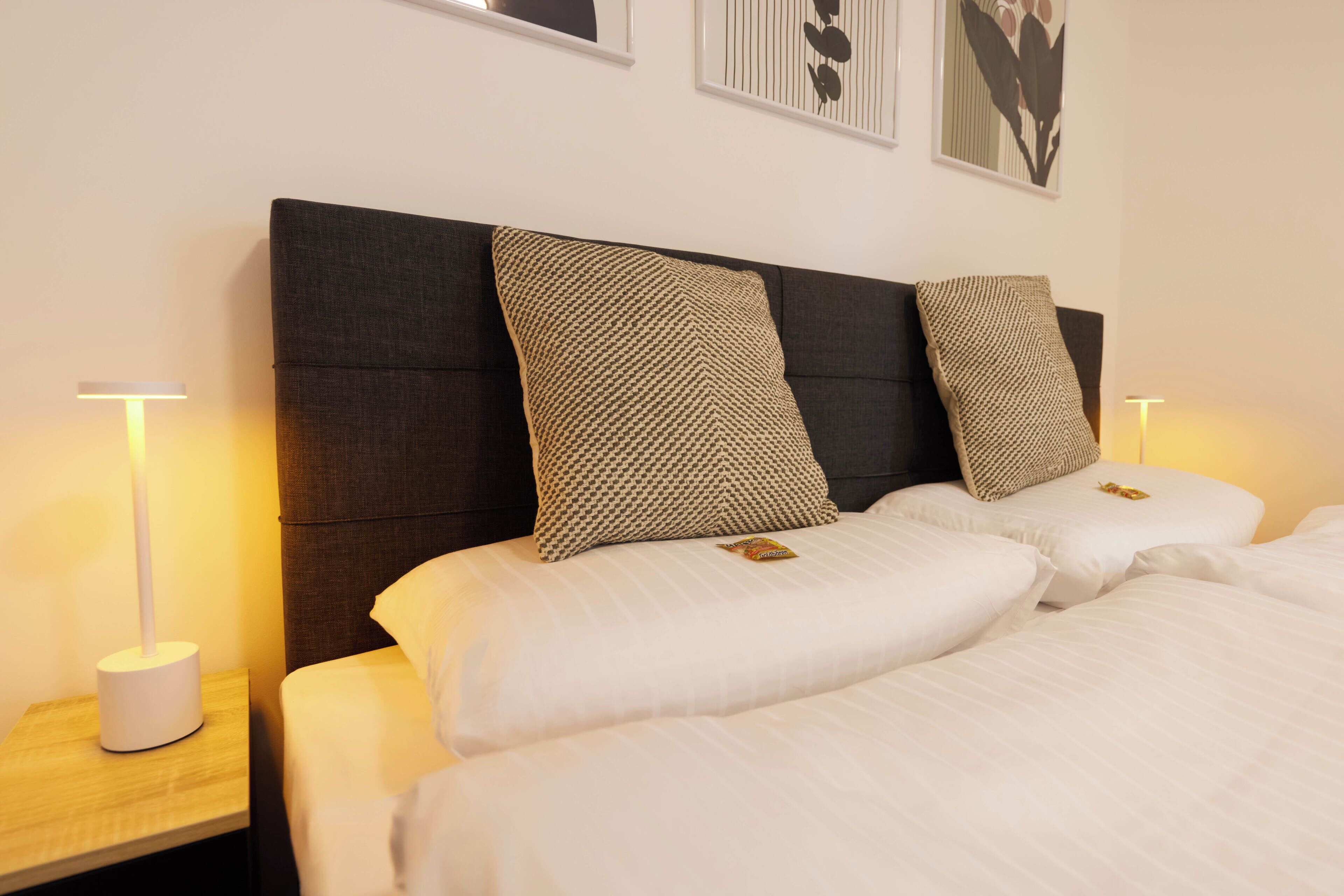 Ein Doppelbett mit zwei Kissen, ein Nachttisch mit Lampe und gerahmte Kunstwerke an der Wand.