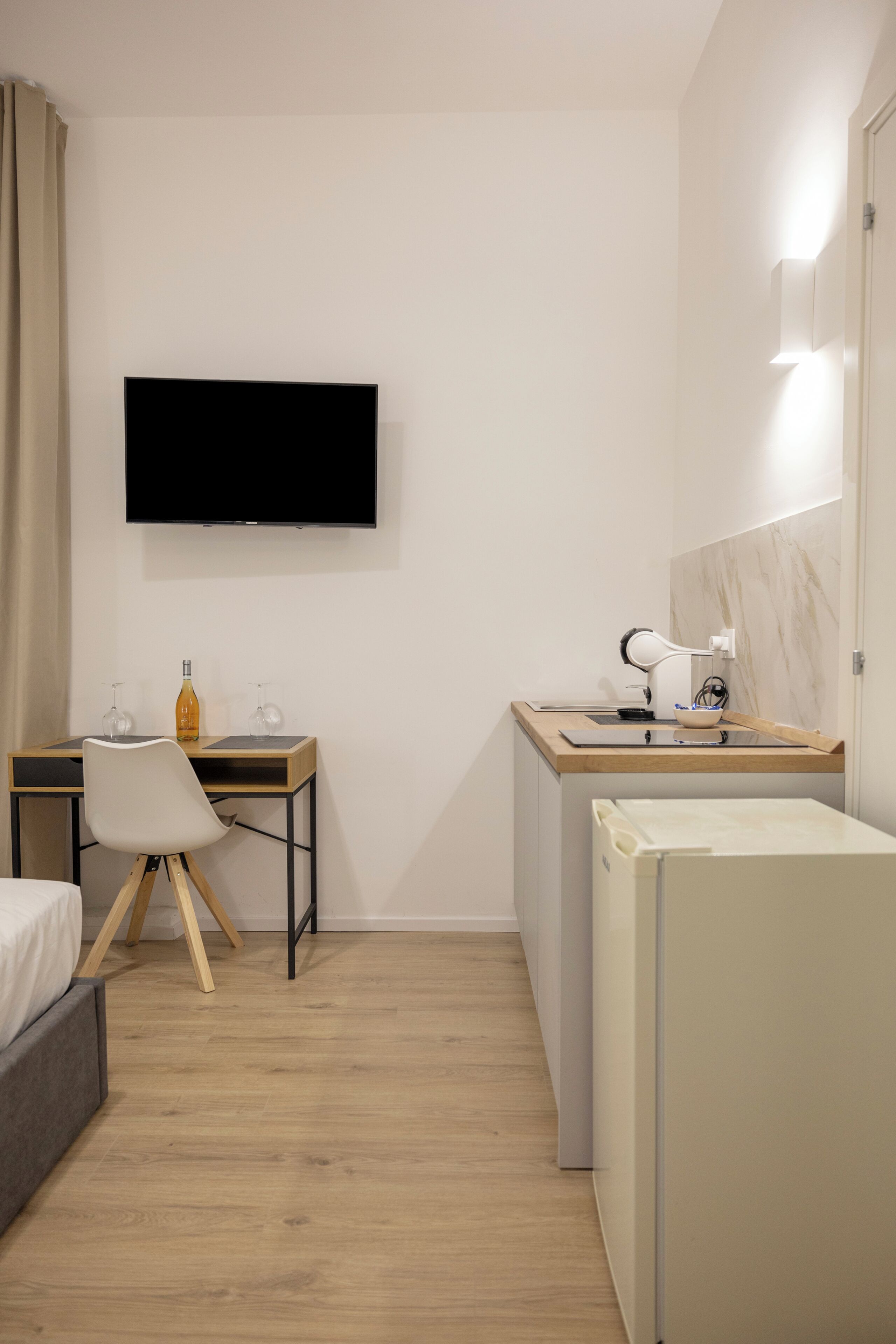 Habitación de hotel pequeña con cama, escritorio y silla, televisor montado en la pared, refrigerador y mesita de noche con lámpara.