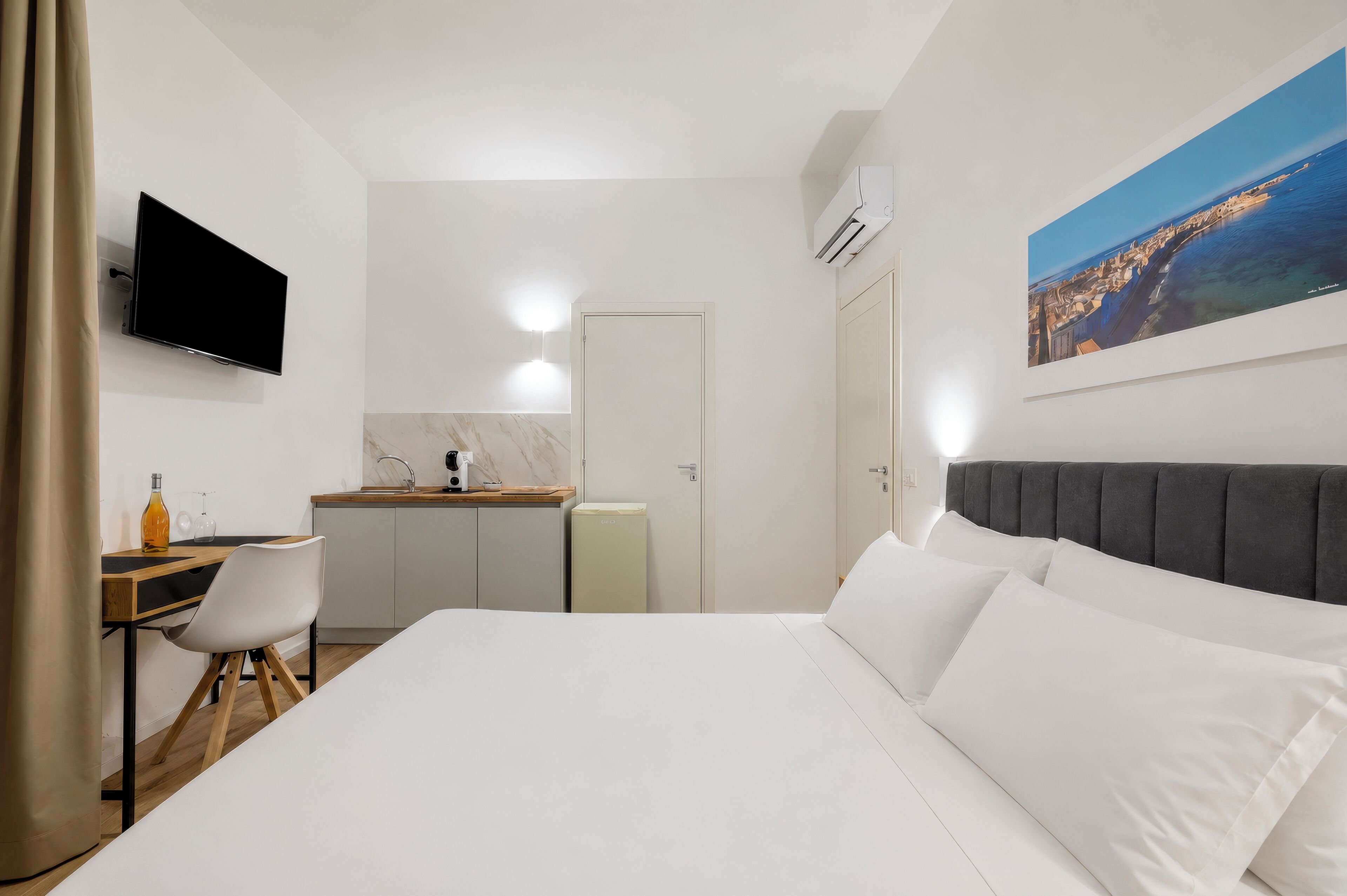 Une chambre d’hôtel comprenant un lit, un bureau avec une chaise, une télévision, un coin cuisine et un tableau au mur.