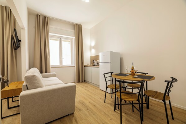 A compact living space with a sofa, a dining table with chairs, a kitchenette, and a window with curtains.