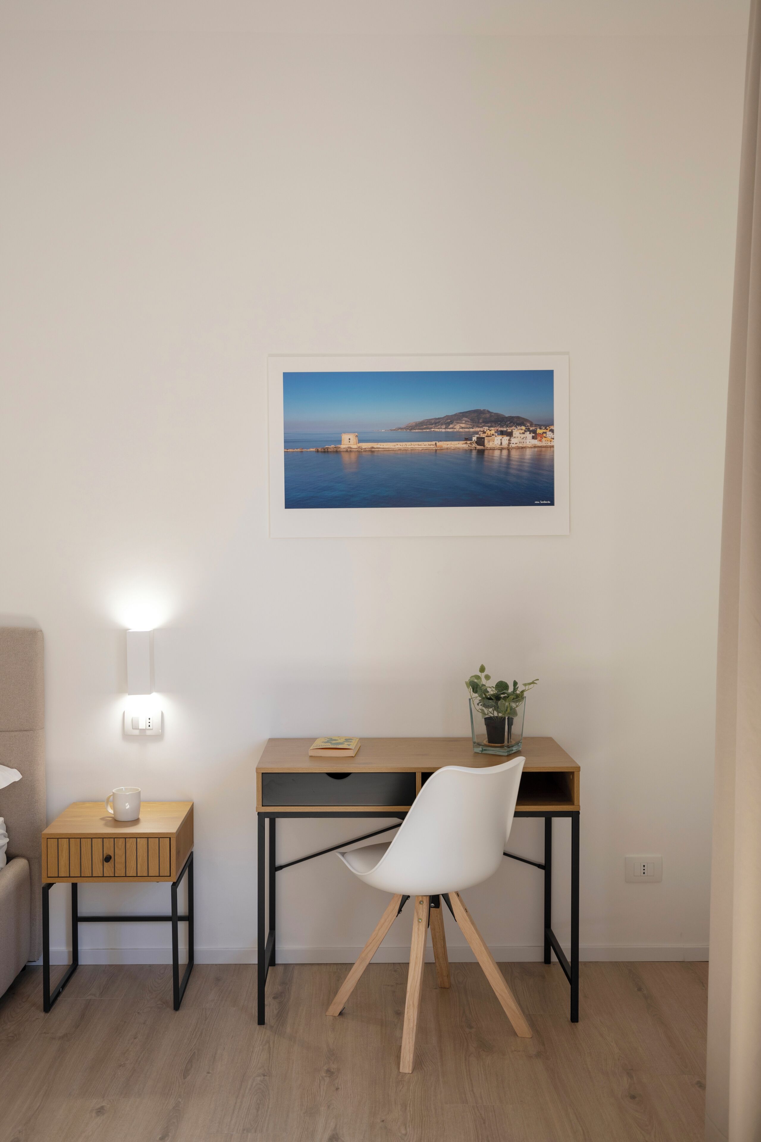 Ett minimalistiskt rum med ett skrivbord i trä, en vit stol, ett litet nattduksbord med en lampa och en inramad tavla med ett kustlandskap på väggen.