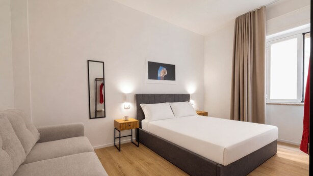 Hotellihuone, jossa on sänky, yöpöytä, seinävalaisin, peili, taulu seinällä ja ikkuna, jossa on verhot.