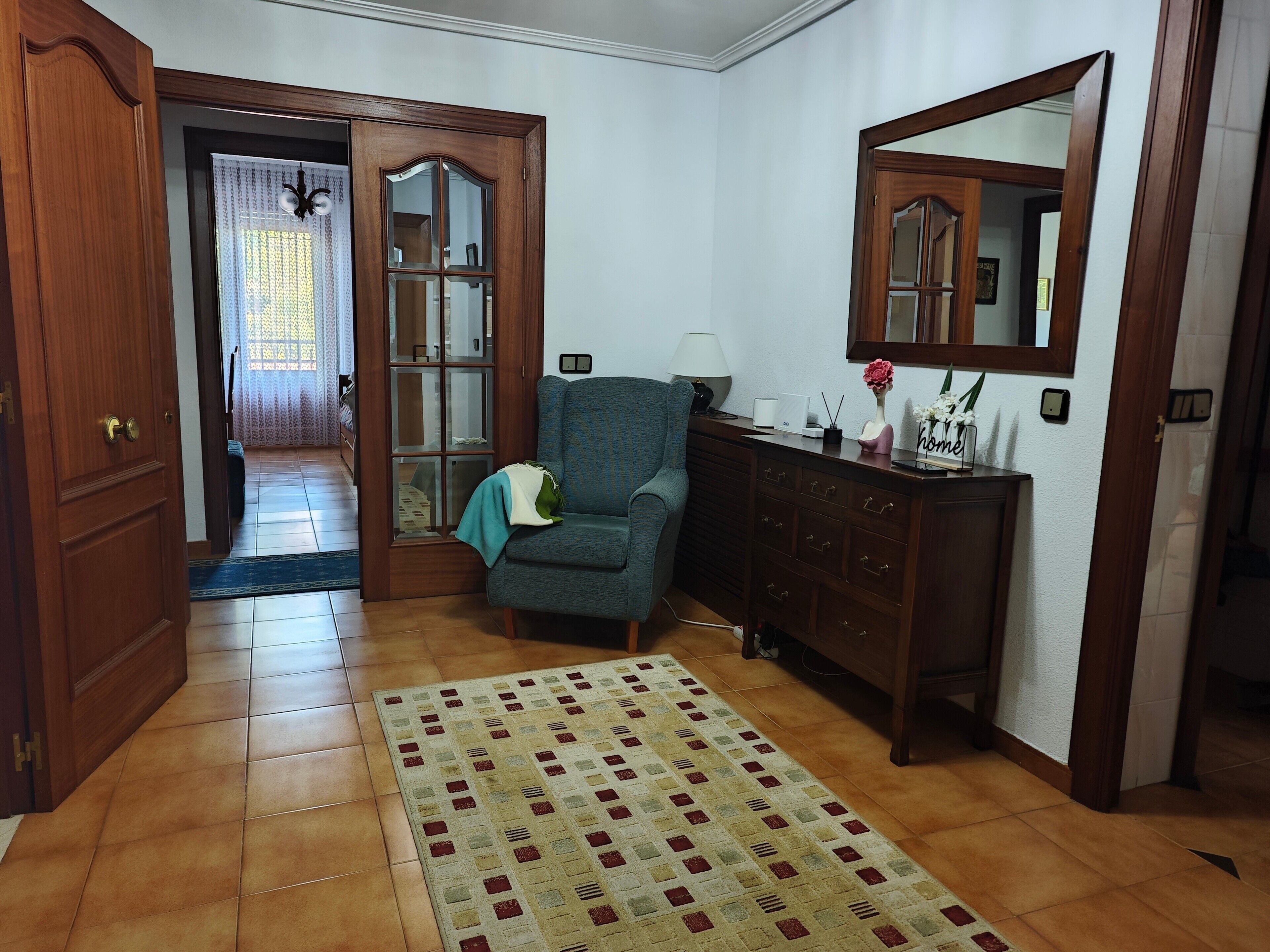 Una habitación con un armario de madera, un sillón azul, un aparador de madera y un espejo en la pared.