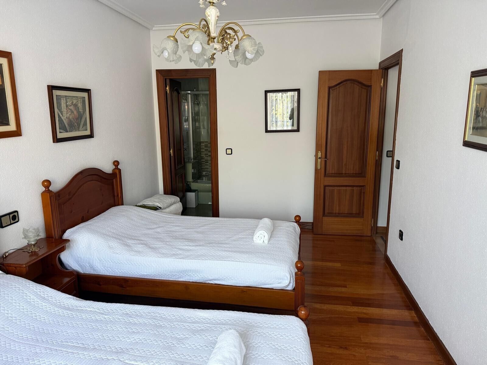A hotel room with two beds, a wooden floor, a chandelier, and framed pictures on the walls.
