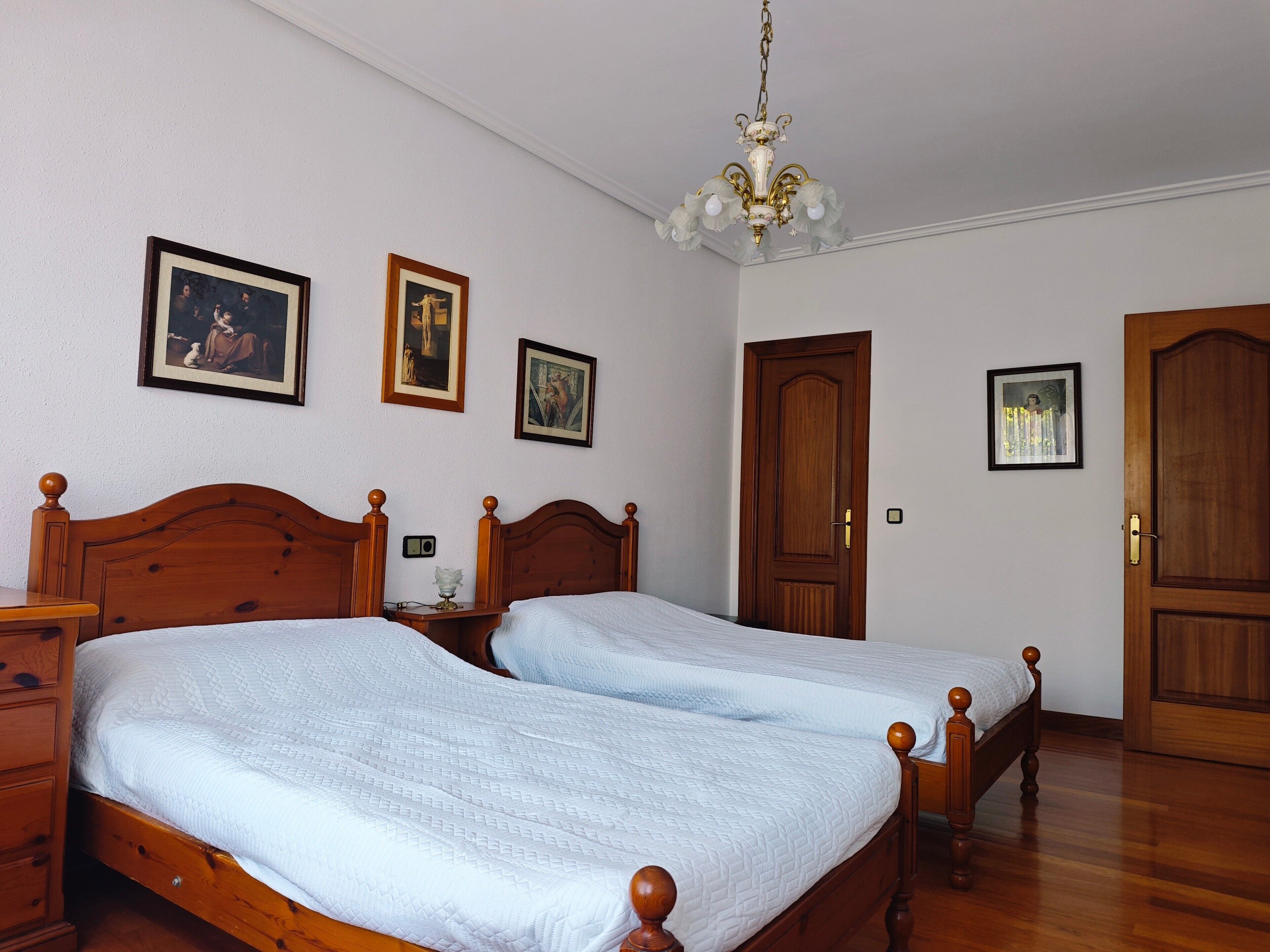 Een slaapkamer met twee houten bedden, een kroonluchter en ingelijste foto’s aan de muur.