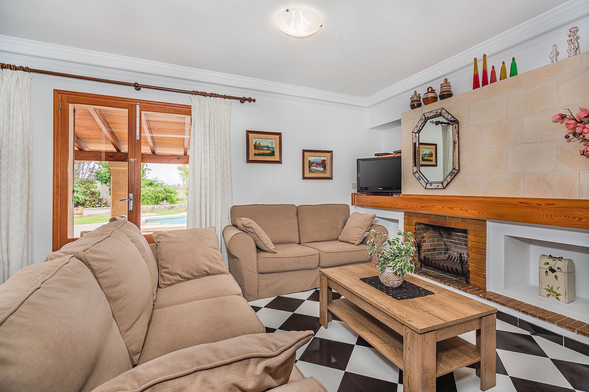 Ein Wohnzimmer mit Schachbrettmuster-Boden, zwei beigen Sofas, einem hölzernen Couchtisch, einem Fernseher und einem Kamin mit Dekoration.