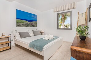 Ein modernes Schlafzimmer mit einem Bett, einem Fenster mit Blick auf Palmen, einem Strandbild und einem Nachttisch aus Holz.