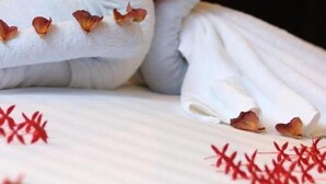 A bed with a heart-shaped towel arrangement and scattered red flowers.