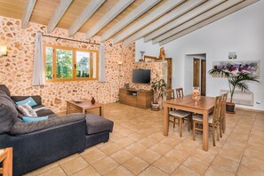 Een woonkamer met een stenen muur, een bank, een televisie en een eethoek met een tafel en stoelen.