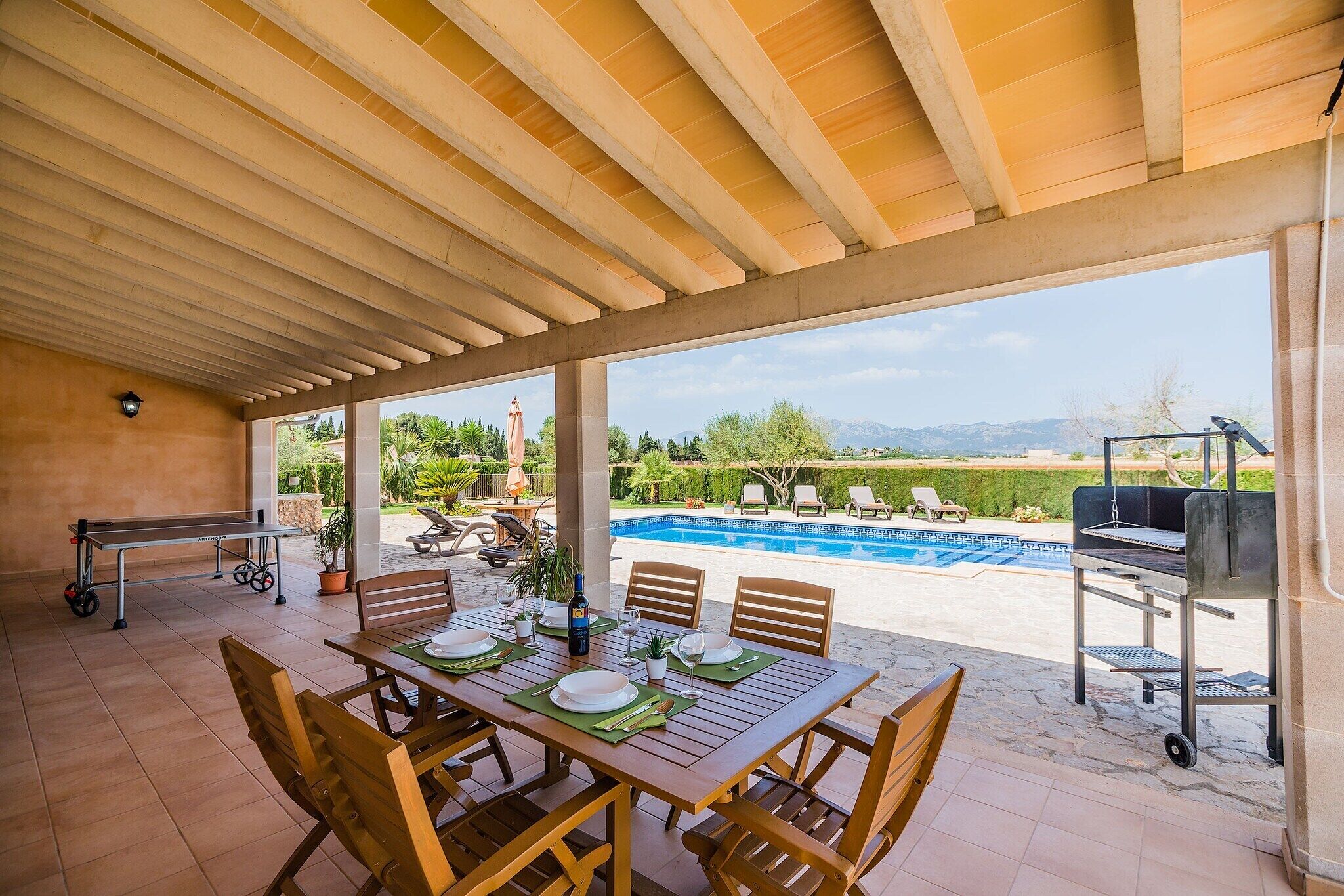 An outdoor patio with a dining table set for four, a barbecue grill, and a pool in the background.