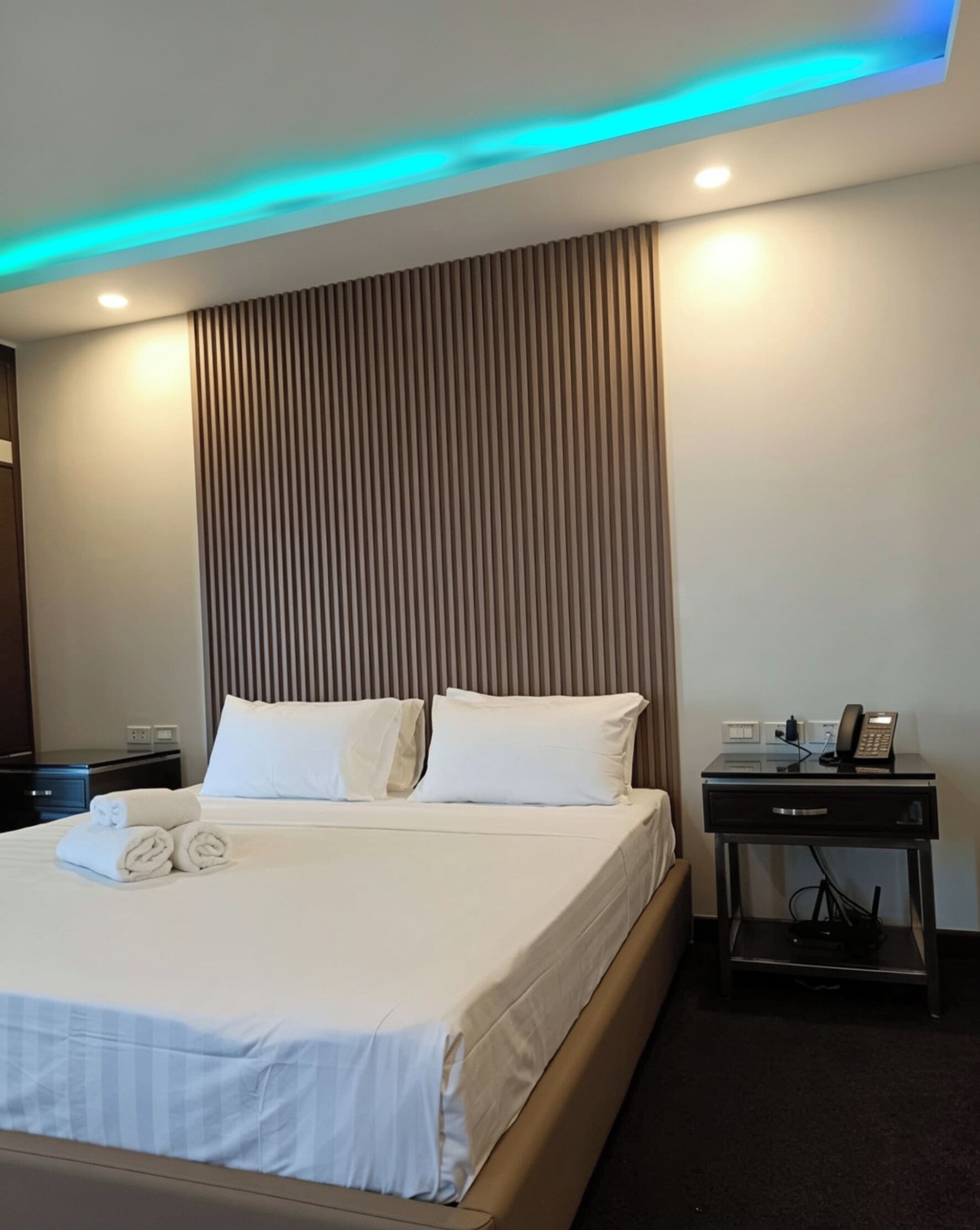 Una habitación de hotel con cama, mesita de noche y teléfono, y una pared con líneas verticales.