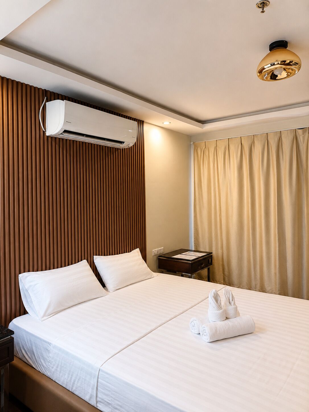 A hotel room with a bed, an air conditioning unit, a nightstand, and a window with curtains.