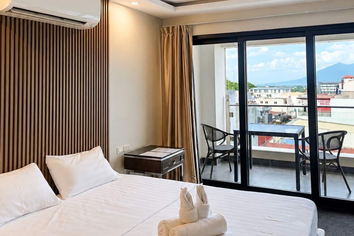 Habitación de hotel con cama, escritorio, silla, equipo de aire acondicionado y balcón con vistas.