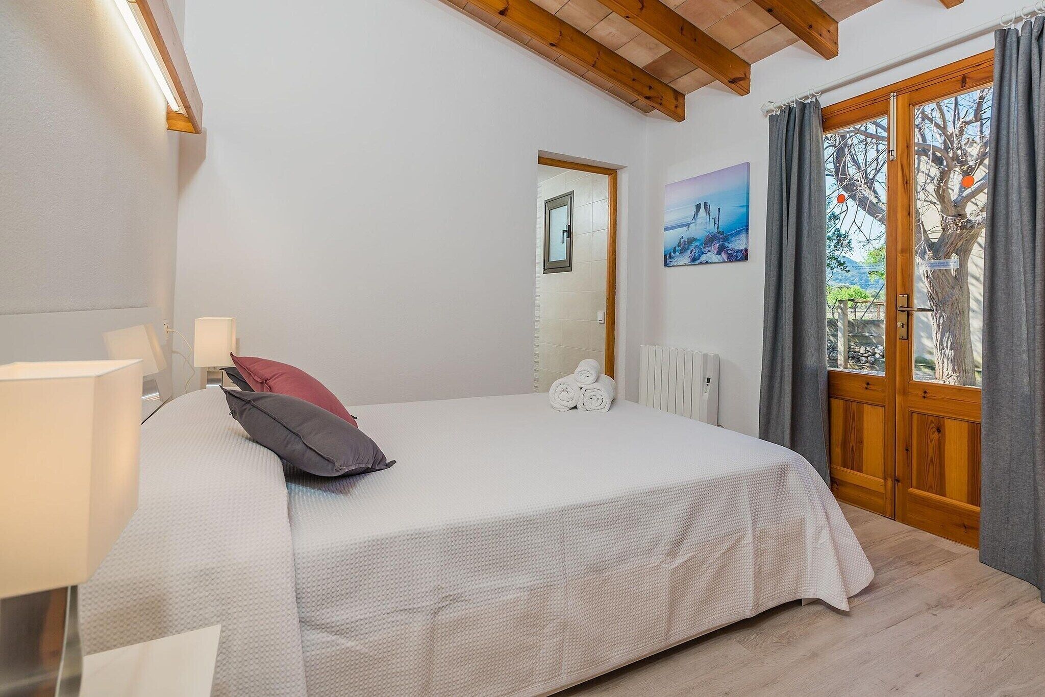 A bedroom with a bed, a nightstand, a lamp, a window with curtains, a radiator, and a painting on the wall.