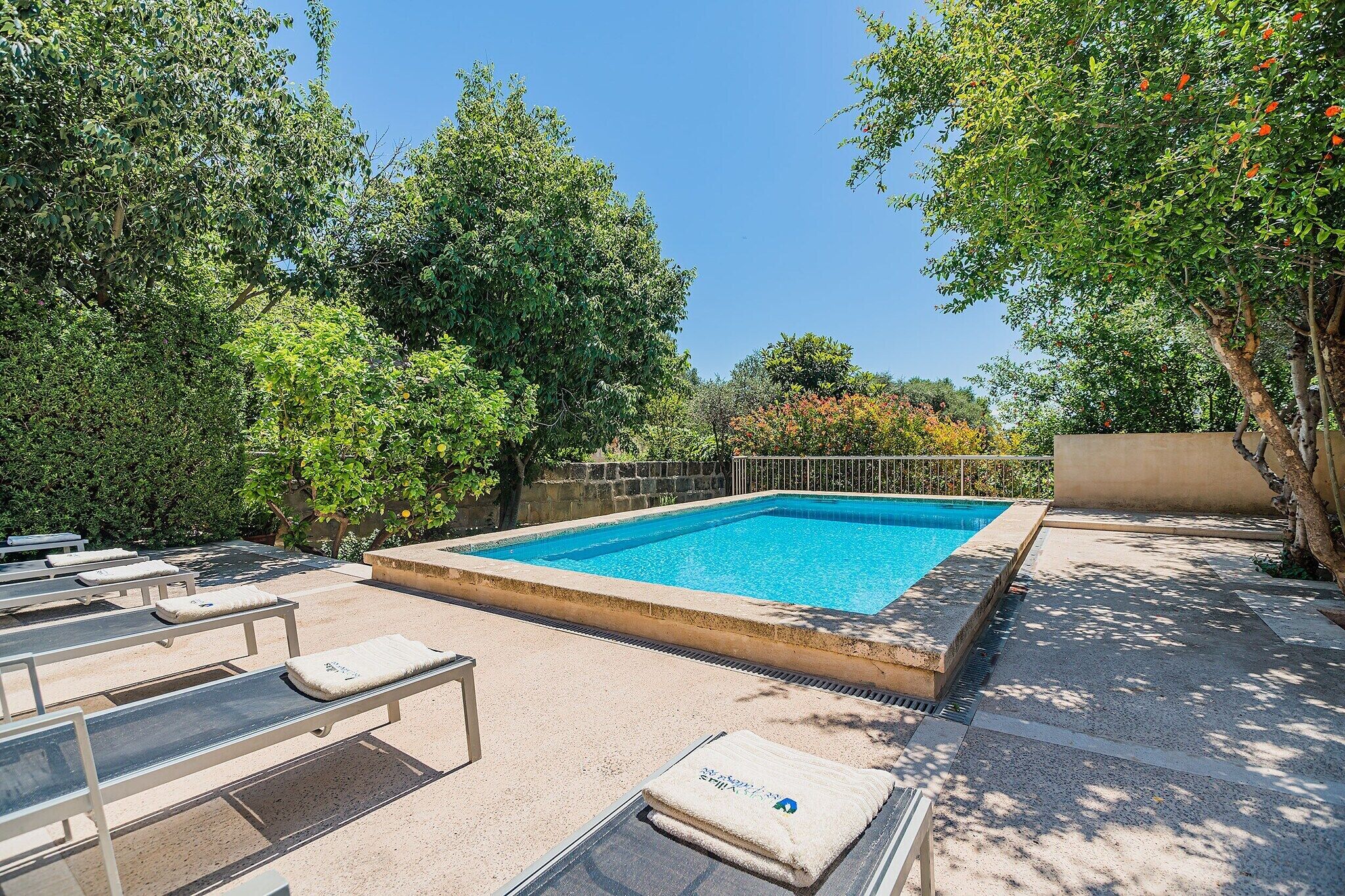 A rectangular swimming pool with a stone border, surrounded by lounge chairs and a stone wall.