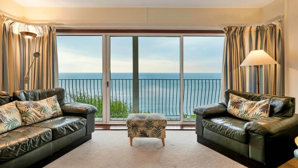 A living room with two leather sofas, a small ottoman, and a large sliding glass door leading to a balcony with a view of the sea. - Lavish Sea-view Holiday Home in Seaton (Torpoint)