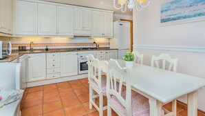 Een keuken met witte kasten, een eettafel met stoelen, een kroonluchter en een schilderij aan de muur.