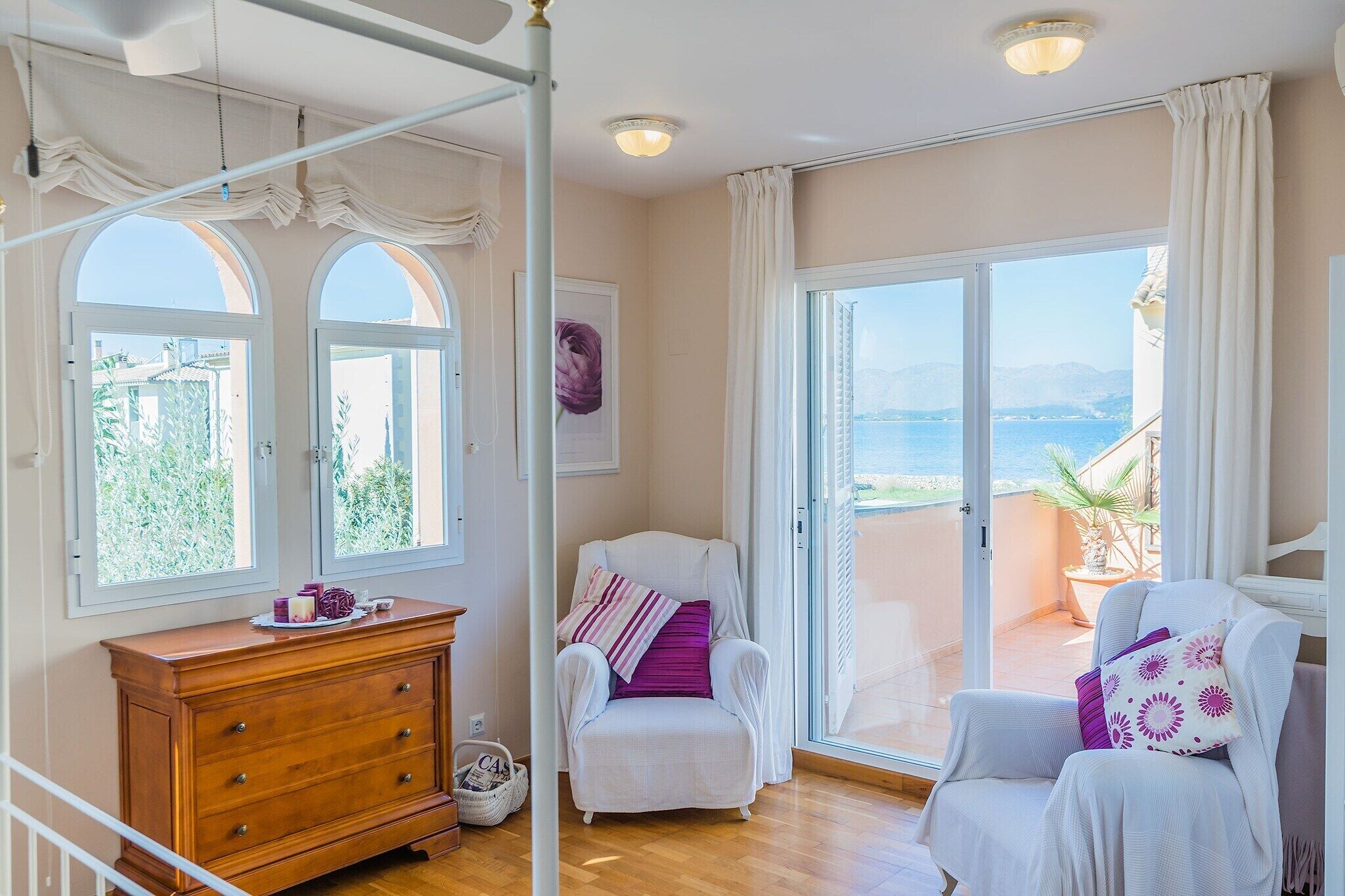 A room with a wooden dresser, a white armchair with a purple pillow, and a view of the sea through a large sliding glass door.